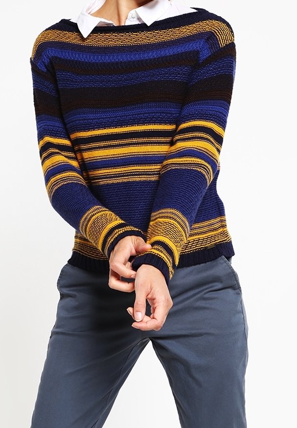 Personne portant un pull en maille rayé marine, bleu, noir et jaune par-dessus une chemise blanche à col et un pantalon gris, ajustant les manches.