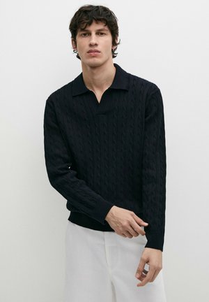 Young man with dark curly hair wearing a black cable-knit sweater and white pants, adjusting his left sleeve against a plain background.