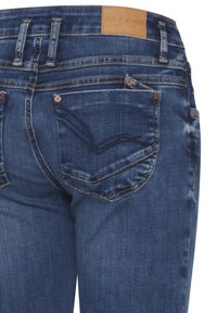 Jeans de mezclilla en azul oscuro con una ligera textura desgastada. Presentan una etiqueta de cuero que dice "PULZ JEANS" y detalles de costura en los bolsillos.