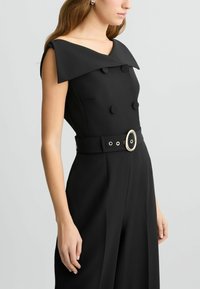Tuta nera con ampio colletto, design doppiopetto e vita aderente con fibbia ovale, texture liscia e vestibilità sartoriale.