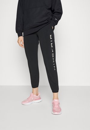 Person wearing black Nike Air sweatpants and pink Nike running shoes, standing against a plain light background.