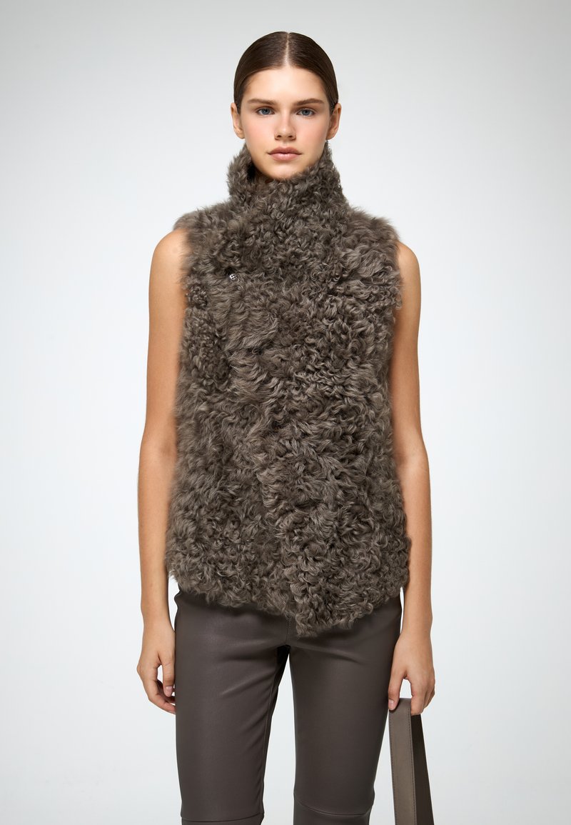 Gilet senza maniche in morbido pelliccia sintetica riccia grigia, con colletto alto e design frontale asimmetrico, abbinato a pantaloni similpelle marroni.