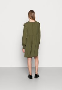 Vihreä öljypuku, jossa on sujuva muotoilu, rypytettyjen olkaimien korostukset ja pitkät hihat. Vaatteen väri on tummanvihreä, ja se ulottuu polven yläpuolelle. Yhdistetty mustiin kenkiin.