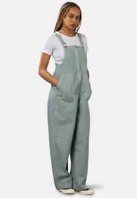 Salopette rayée bleu et blanc avec jambes larges, fermeture éclair à l'avant, bretelles réglables et grandes poches. Portée par-dessus un t-shirt blanc.