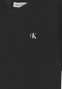 Czarna sukienka z gładkiego materiału, z okrągłym dekoltem i białym logo "ck" po lewej stronie. Prosty, dopasowany krój.