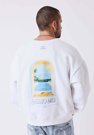 Uomo con capelli corti e collana a catena indossa una felpa bianca con un disegno colorato a finestra che mostra piscina, oceano e testo su Monaco.