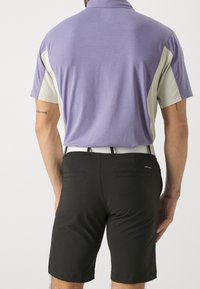 Homme portant un short noir avec des poches arrière et une chemise polo à manches courtes violette et beige, se tenant de dos face à l'appareil photo.