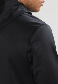 Sudadera negra con una textura satinada suave, que presenta una capucha redondeada y un diseño ajustado. Costuras visibles a lo largo de la costura del hombro.