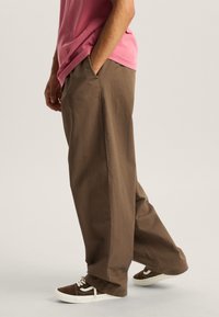 Brown wide-leg pants made of cotton, featuring side pockets and a smooth texture. Worn with dark brown sneakers and a pink shirt.