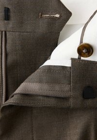 Gros plan de la ceinture d'un pantalon marron à motif pied-de-poule montrant un bouton, une fermeture éclair, un crochet de fermeture et une doublure intérieure blanche avec des coutures marron.