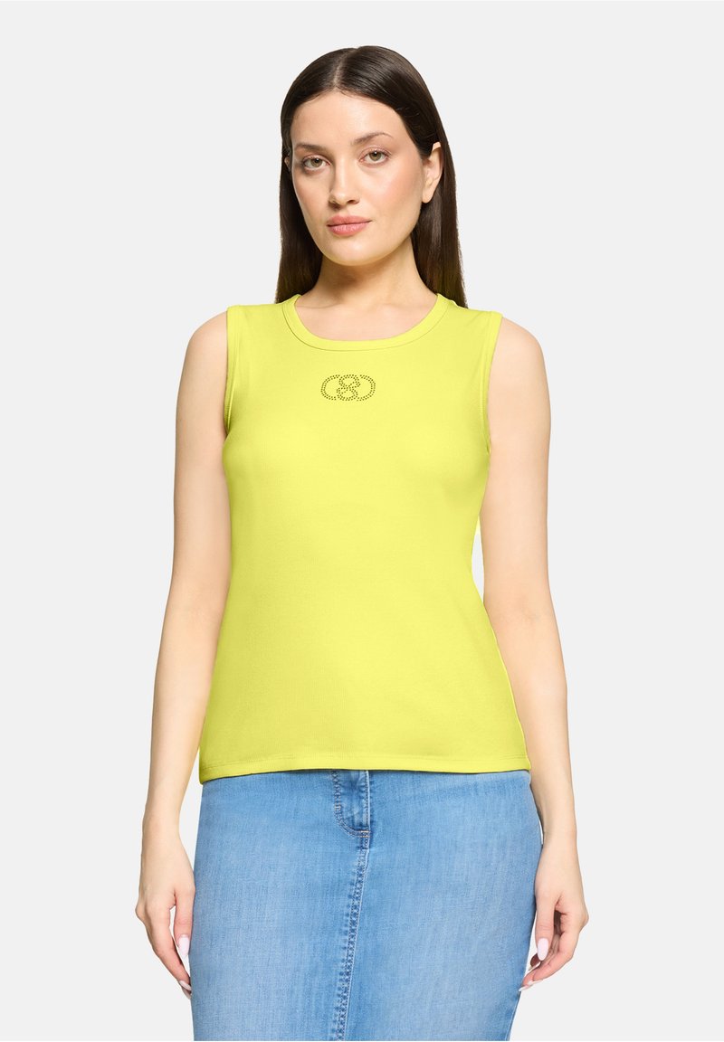 Femme aux cheveux bruns et raides portant un haut sans manches vert citron avec un petit logo et une jupe en jean bleu clair, debout devant un fond blanc.