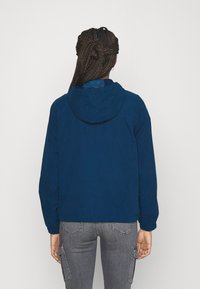 Osoba z warkoczami, ubrana w niebieską kurtkę z kapturem i szare dżinsy cargo, stojąca tyłem na tle jasnym.