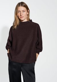 Maglione marrone scuro, testurizzato, con un collo alto e arrotondato e spalle cadenti; abbinato a pantaloni neri lisci con tasche laterali.