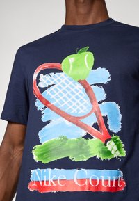 Granatowa bawełniana koszulka z grafiką rakiety tenisowej z jabłkiem, malowanymi teksturami w zieleni i błękicie oraz napisem "Nike Court".