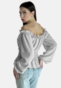 Top grigio chiaro fuori spalle con scollatura arricciata e vita smock, caratterizzato da maniche lunghe e morbide e una texture liscia. Abbinato a jeans chiari.
