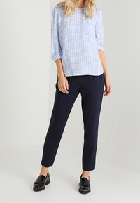 Himmelbluse mit weißem Karo-Muster, langen Ärmeln und einer Tasche auf der linken Brust, kombiniert mit dunkelblauen Slim-Fit-Hosen und schwarzen Loafers.