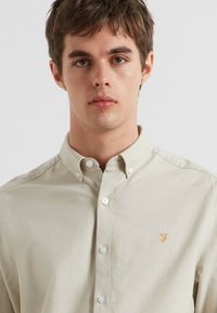 Beige overhemdbloes met kraag gemaakt van katoen, met een klein geborduurd logo in oranje en witte knopen.