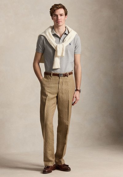 Jaunas vyras stovi su rankomis kišenėse, dėvi pilką polo marškinėlį, smėlio spalvos kelnes, baltą megztinį per petį ir rudus loaferius.