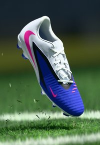 Λευκό, μπλε και ροζ παπούτσι ποδοσφαίρου Nike στον αέρα πάνω από το γρασίδι με σκορπισμένη βρωμιά, που δείχνει δυναμική κίνηση στο γήπεδο.