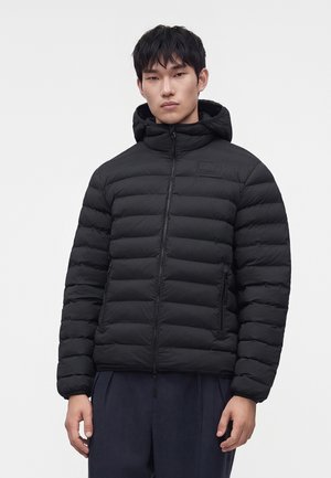 Jeune homme portant une veste matelassée noire à capuche avec poches zippées et pantalon foncé, debout devant un fond clair uni.