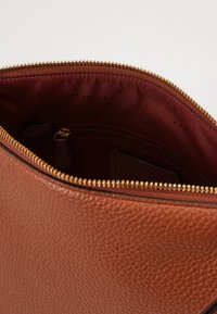 Bolsa de couro castanho texturizado com fecho éclair. O interior apresenta um forro bordô rico, um bolso com fecho e slots para cartões.