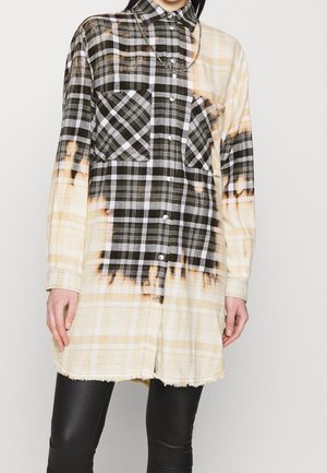 Femme portant une longue chemise boutonnée à carreaux bicolore noir et beige avec poches poitrine et leggings noirs, montrant un collier chaîne et un ourlet effiloché.