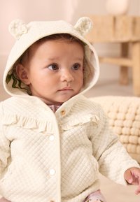 Veste à capuche matelassée crème avec oreilles d'ours, volants sur le devant, boutons dorés, et doublure fleurie. Texture douce.