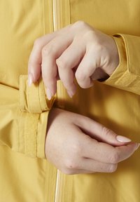 Żółta kurtka o gładkiej strukturze, z dopasowanym mankietem zabezpieczonym paskiem na rzep. Zbliżenie rąk regulujących mankiet.