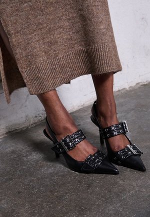 Piernas de piel oscura con tacones negros de punta afilada con hebillas y tiras plateadas, combinados con una falda midi de punto marrón sobre un suelo de hormigón.