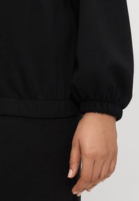 Svart hettegenser med elastiske mansjetter og nederkant. Glatt tekstur med et enkelt design, som primært viser armen og håndleddet.