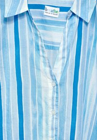 Camisa a rayas azul y blanca hecha de un tejido ligero. Presenta un escote en pico y un cierre de botones con una etiqueta que dice "respira".