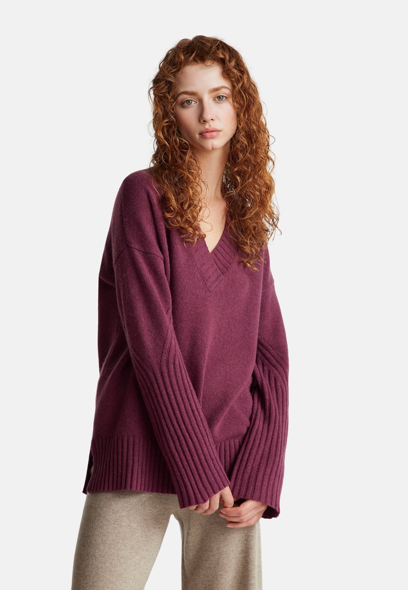 Übergroßer, V-Ausschnitt-Pullover in tiefem Pflaumenfarben mit gerippten Ärmeln und Saum. Weiche Textur, seitliche Schlitze für Bewegungsfreiheit, kombiniert mit hellbeigen Hosen.