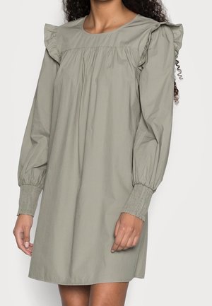 Femme portant une robe ample à manches longues de couleur vert sauge avec des détails froncés aux épaules et des poignets smockés.