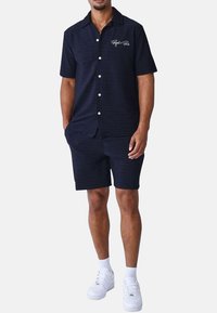 Camisa de botones de manga corta color marino con rayas horizontales y bolsillo frontal, combinada con pantalones cortos a juego, usada con zapatillas blancas.