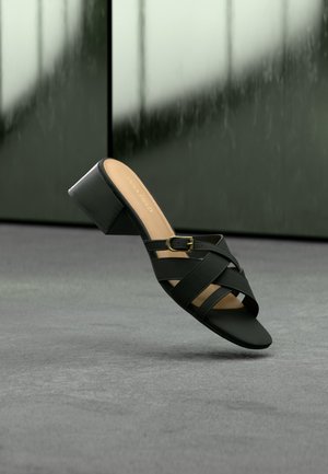 Μαύρο σανδάλι με τακούνι, με διασταυρούμενους ιμάντες και λεπτομέρεια με χρυσή αγκράφα, σε φόντο με θαμπό σκουροπράσινο και γκρι χρώμα.