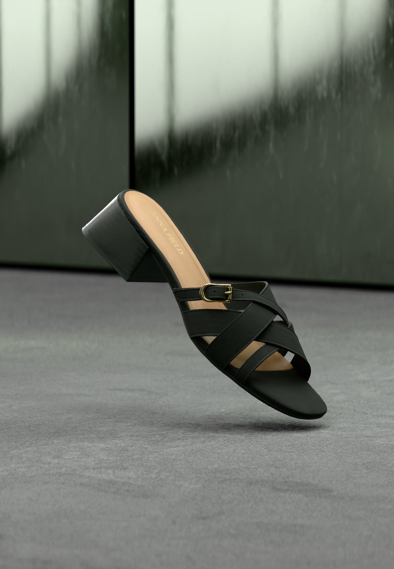 Sandale à talon noire avec brides croisées et détail boucle dorée, présentée sur un fond flou vert foncé et gris.