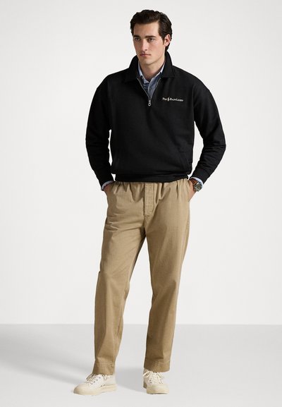 Pulover negru cu fermoar pe jumătate și logo, asociat cu pantaloni bej. Modelul poartă adidași de culoare deschisă și o brățară. Design simplu și casual.