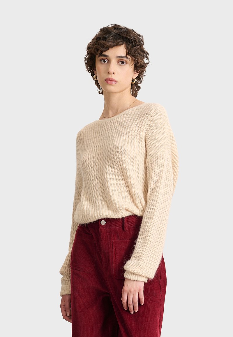 Suéter de punto beige con amplio escote barco, corte holgado y mangas largas, combinado con pantalones de pana burdeos de talle alto que cuentan con bolsillos delanteros.