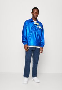 Blå nylon genserjakke med halv glidelås, med "USA"-logo i gult, kombinert med blå jeans og hvite joggesko.