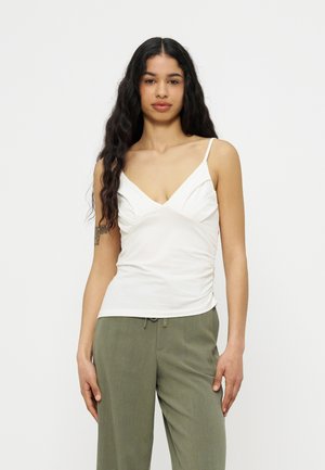 Kobieta z długimi czarnymi włosami, ubrana w białą, bezrękawnikową bluzkę z dekoltem w serek oraz oliwkowe spodnie z sznurkiem, na jednolitym tle.