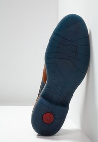 Scarpa in pelle marrone con una suola in gomma blu texture. Presenta un logo rosso tondeggiante sul tallone e perforazioni decorative sul lato.