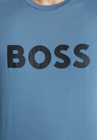 Kék pamut póló kerek nyakkivágással, amelyen vastag fekete "BOSS" felirat található a mellkason. Simább textúrájú, laza dizájn.