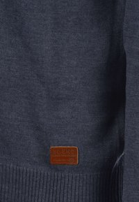 Mörkblå stickad tröja med ribbad nederkant och ärmslut. Har en brun läderetikett med texten "BLEND GLOBAL JEANMAKER" präglad.