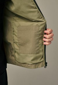 Veste vert olive avec une texture lisse, dotée d'une fermeture éclair, avec un côté ouvert révélant une poche sur la doublure intérieure.
