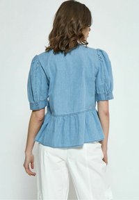 Top peplum in denim bluastro con maniche a sbuffo, polsini abbottonati e cucitura in vita arricciata, mostrato da dietro su uno sfondo neutro.
