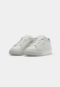 Sneakers bianchi a basso profilo di Tommy Hilfiger, con lacci, suola in gomma e un piccolo logo sul lato, mostrati su uno sfondo bianco semplice.