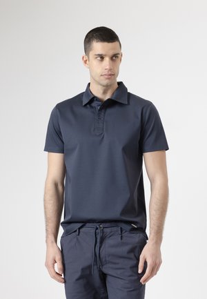 Hombre vistiendo una camiseta polo azul marino oscura y pantalones con cordón a juego, posando contra un fondo liso de color gris claro, mirando hacia un lado.