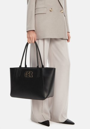 Persona vestida con traje y pantalones beige, sosteniendo un bolso tote de cuero negro con logo dorado y doble asa, calzando zapatos negros puntiagudos.