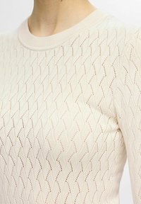 Ljust beige stickad tröja med en vågig mönstring, som har en rund halsringning och strukturerad tyg, med öppna maskor genom hela plagget.