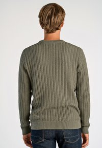 Pull olive en tricot avec une texture c�ôtelée, encolure ronde et manches longues. Présente une taille et des poignets ajustés. Porté avec un jeans foncé.